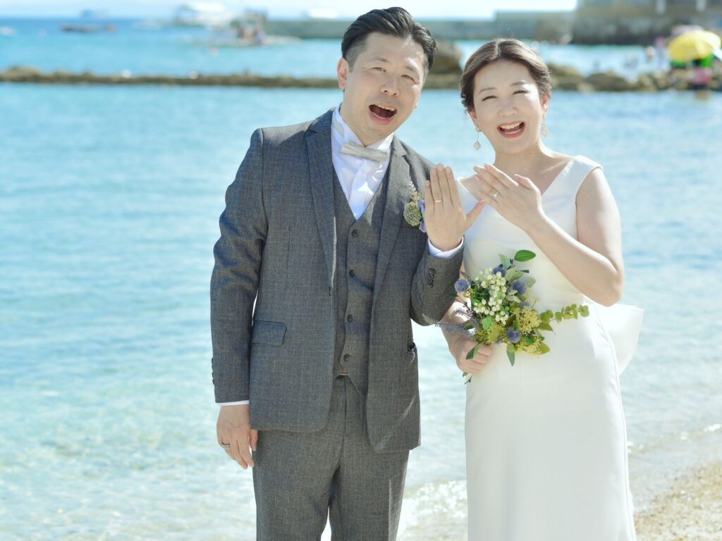 海を背景に結婚指輪を見せながら笑顔の新郎新婦の前撮りフォト